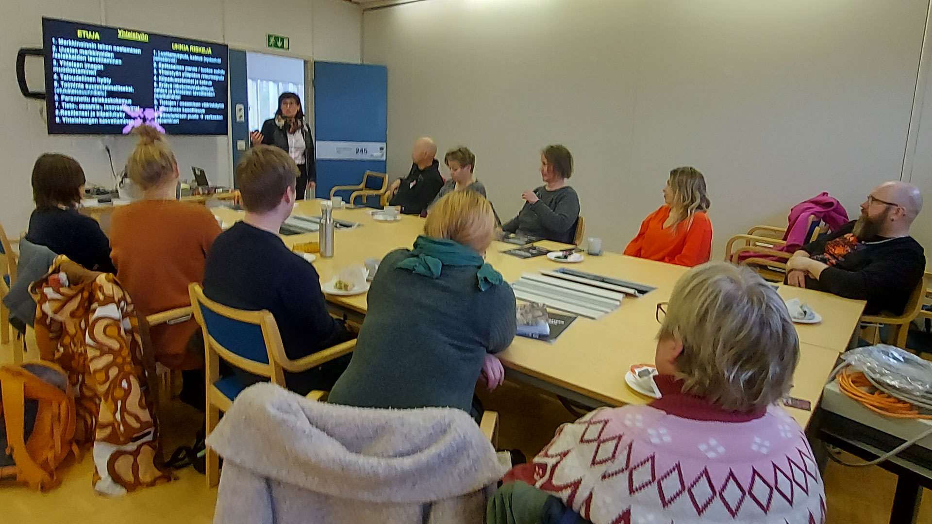 Ihmisiä pöydän ääressä seuraamassa kouluttajan pitämää esitystä.