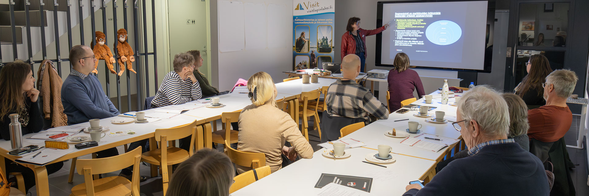 Ihmisiä pöytien ääressä katsomassa huoneen etualalla olevaa kouluttajaa, joka osoittaa valkokankaalla näkyvää esitystä.