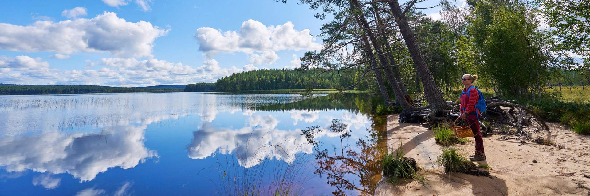 Nainen seisoo hiekkarannalla katsoen järvelle. Pilvet heijastuvat järven pinnasta.