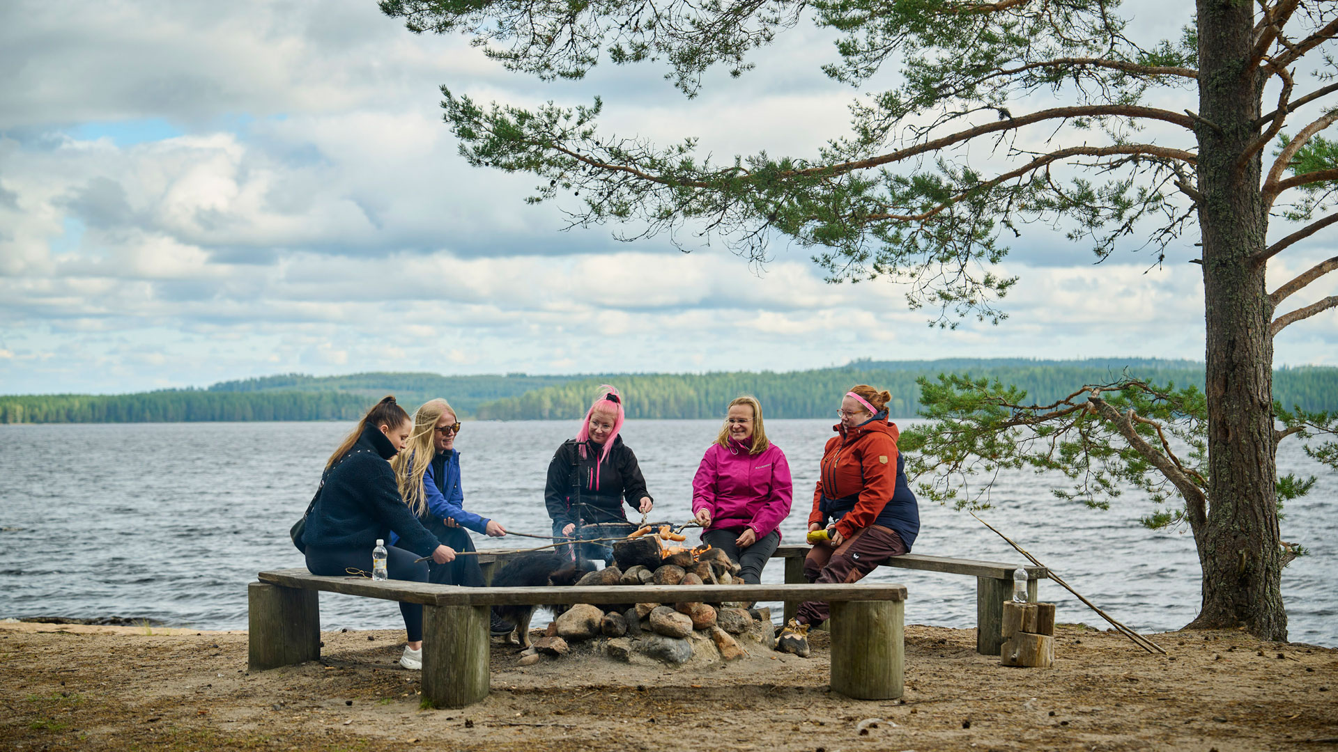 Viisi naista istuu nuotion äärellä hiekkarannalla ja paistaa makkaraa. Taustalla on järvi.