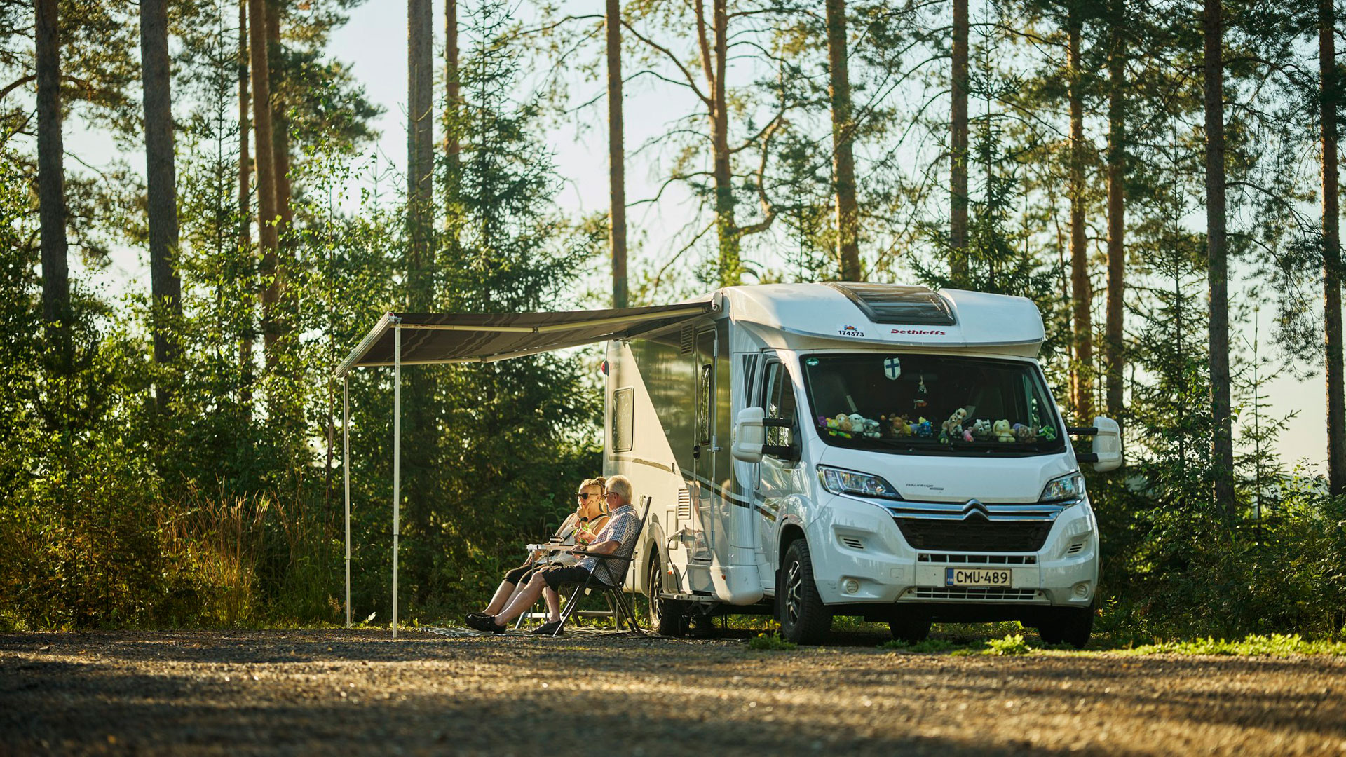 Matkailuauto parkissa, jonka sivukatoksen alla istuu retkituoleilla kaksi henkilöä.